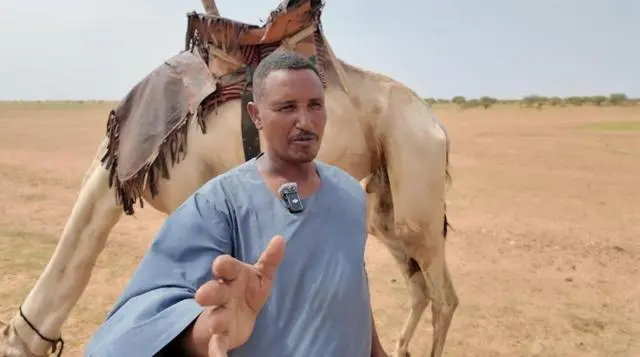 رجل في الأربعينات من عمره يتحدث وخلفه جملاً يرعى.