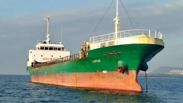  تظهر السفينة "إم في لُطف" راسية بالقرب من الساحل، ويمكن رؤية هيكلها الأخضر والبرتقالي فوق سطح الماء.
