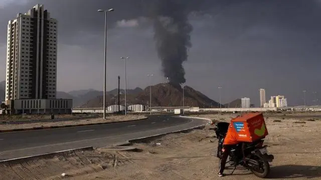  شخص يقف بجوار دراجة نارية بينما يتصاعد الدخان في منطقة صناعة النفط بالفجيرة، الإمارات، إثر حريق نجم عن حطام بعد اعتراض الدفاعات الجوية لطائرة مسيرة،  مارس/آذار.