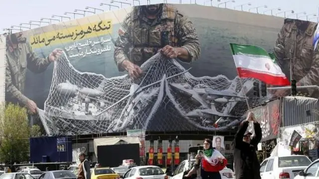  لافتة كبيرة معلقة في شارع في  طهران كتب عليها: "سيبقى مضيق هرمز مغلقاً؛ والخليج بأكمله هو ساحة صيد لنا".
رجل يحمل العلم الإيراني وسيدة ترتدي وشاح العلم الإيراني وتحمل صورة المرشد مجتبة الخامنئي